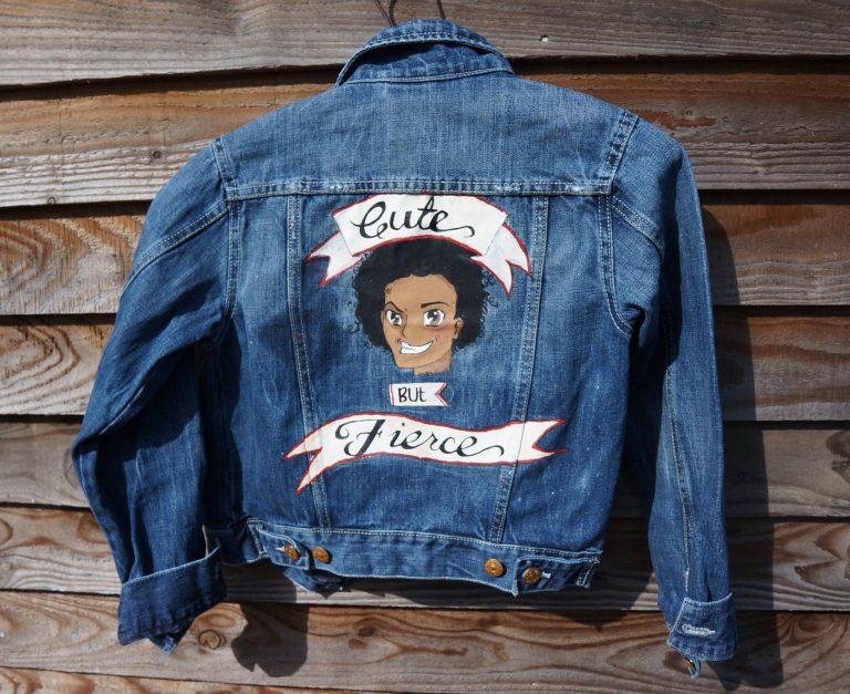 childs denim jacket. It pictures a cartoon image of a brown skinned boy with big curly hair and feirce smile. above the image is the word "cute" on a banner, and below, "but fierce" on another banner.
