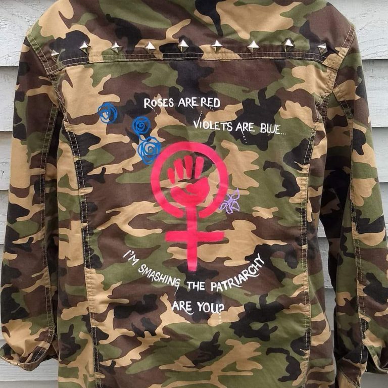 the rear side of a camoflage jacket. it has studs across the shoulders. In the middle is a hot pink feminist symbol with the female sign plus a clenched fist inside the circle. The words "roses are red, violets are blue, I'm sashing the patriarchy, are you?" is written in white