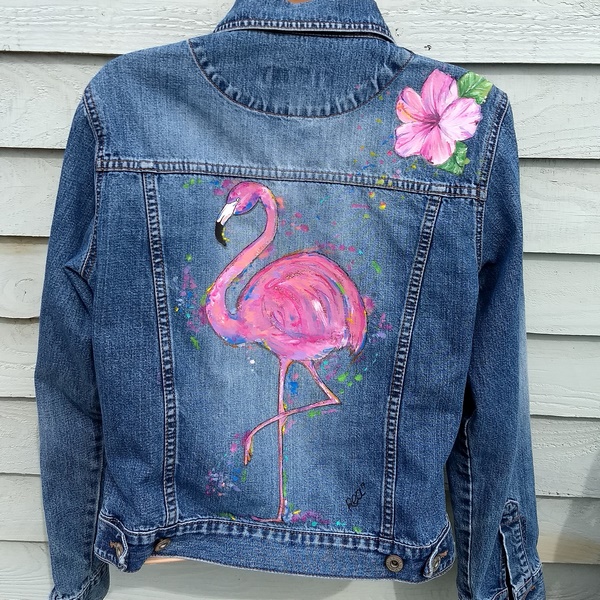 the back of a denim jacket with the picture of a famingo on it surrounded by paint splats. on the right hand shoulder is a hibiscus flower.