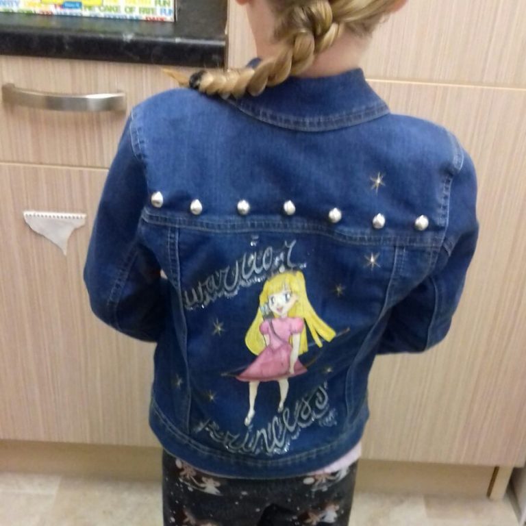 the back of a girl with a blonde plaited pony tail that has been pushed ton the left side to reveal the back of her denim jacket. Across the shoulders of the jaket is a row of round studs. Below the shoulders is a picture of a cartoon princess with long blonde hair and a pink dress. SHe is carrying a bow and arrow. the words "warrior princess are written in cursive handwriting.