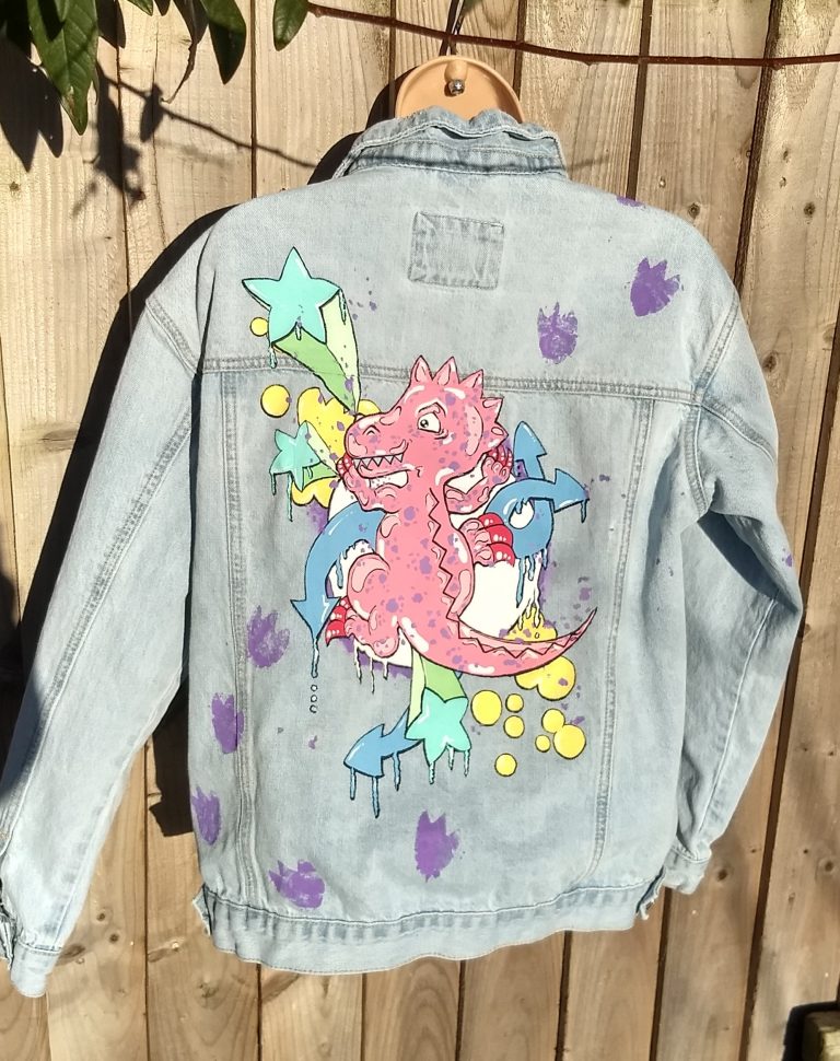 denim jacket with a cute picture of a pink dinosaur climbing up it. There are purple dinosaur prints travelling up from the left hip to the right shoulder. Pictured behind the dinosaur are various graffiti style stars, arrows and bubbles. The theme colour is pastel